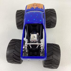 Op de afbeelding: Een blauw monstertruckspeelgoed met grote zwarte banden. De truck heeft een gedetailleerde motor en een dakontwerp met een goud- en bruinpatroon. Blauwe sterstickers versieren de bovenkant van de truck.
