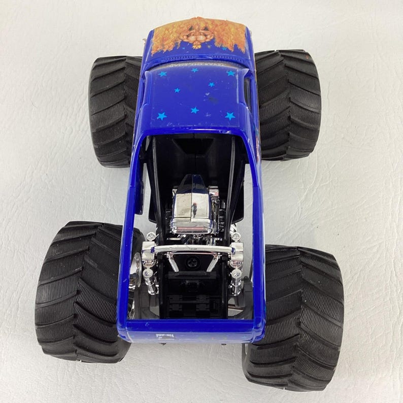 Op de afbeelding: Een blauw monstertruckspeelgoed met grote zwarte banden. De truck heeft een gedetailleerde motor en een dakontwerp met een goud- en bruinpatroon. Blauwe sterstickers versieren de bovenkant van de truck.