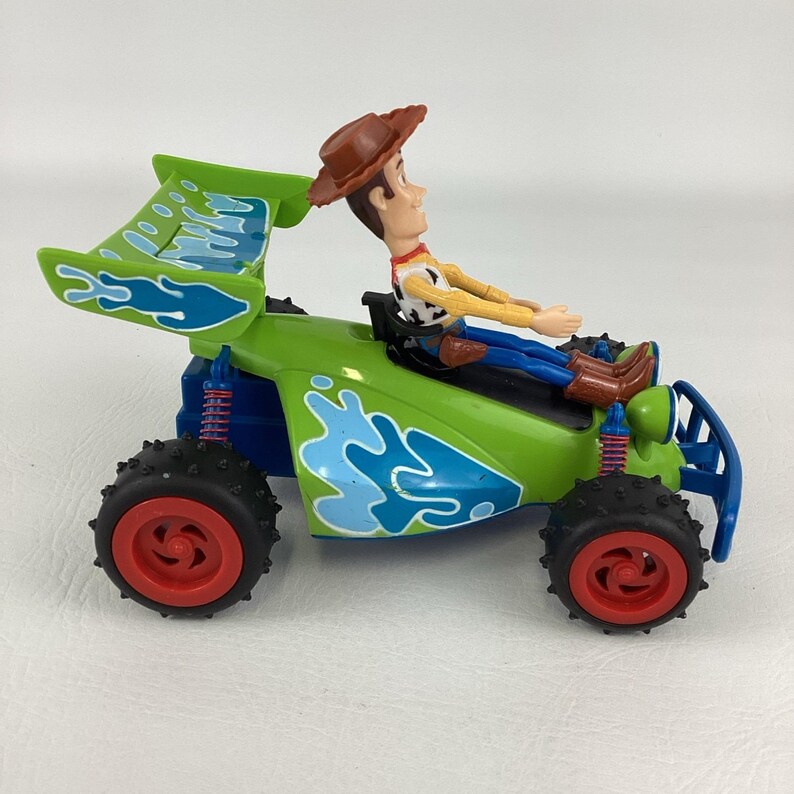 May include: A green and blue toy race car with a Woody figure in the driver's seat. The car has red wheels and a large spoiler. The Woody figure is wearing a cowboy hat, yellow shirt, and blue pants.
