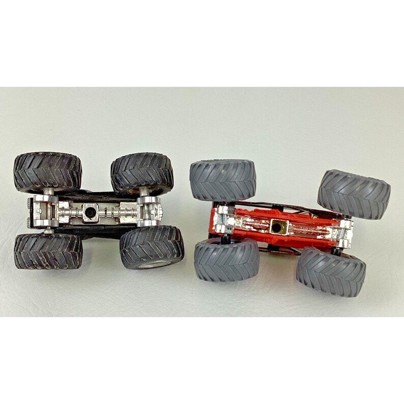 May include: Two toy monster trucks, one black and one red, are shown upside down. The black truck has black tires, and the red truck has gray tires. Both trucks have silver chassis and large tires.