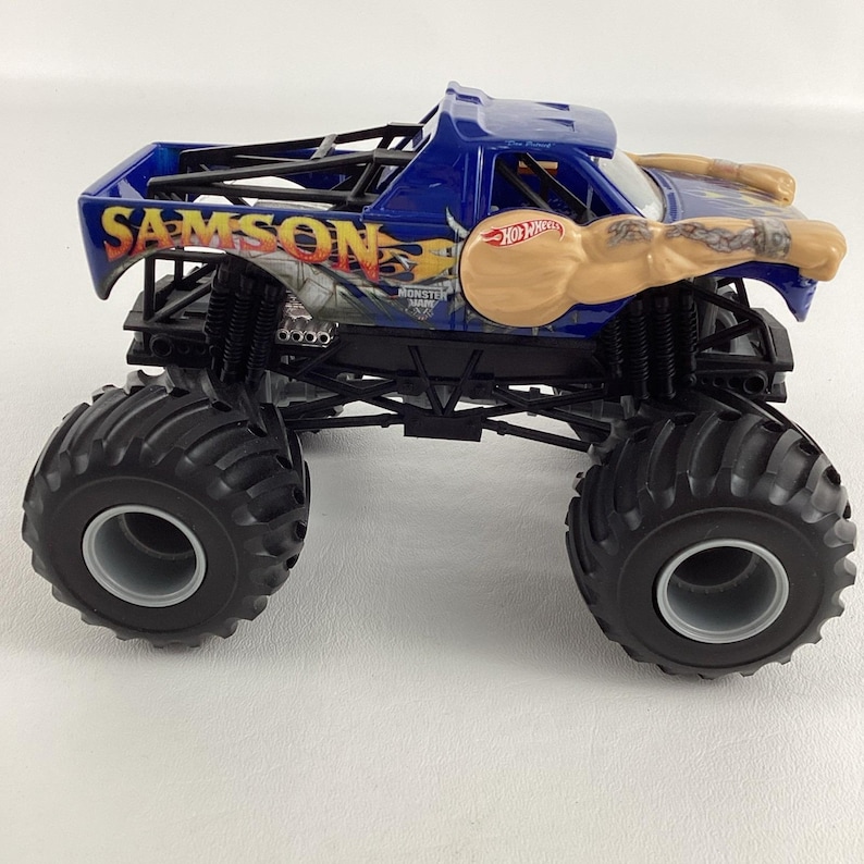 May include: A blue Hot Wheels Samson monster truck toy. The truck features large black tires, a blue cab, and the name "SAMSON" in yellow lettering. The truck has a graphic of a muscular arm and the Hot Wheels logo.