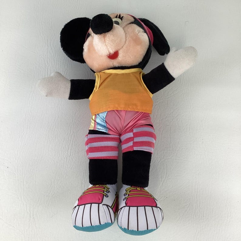 May include: A plush Minnie Mouse doll featuring a yellow sleeveless top, pink shorts with light blue accents, and pink and white striped leg warmers. The doll wears pink and white sneakers with yellow laces and has black ears and white gloves.