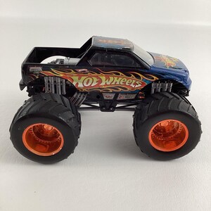 Puede incluir: Un juguete de cami&oacute;n monstruo Hot Wheels azul y negro con gr&aacute;ficos de llamas naranjas y amarillas. El cami&oacute;n tiene neum&aacute;ticos negros grandes con llantas naranjas. El logotipo de Hot Wheels se muestra de forma destacada en el lateral.
