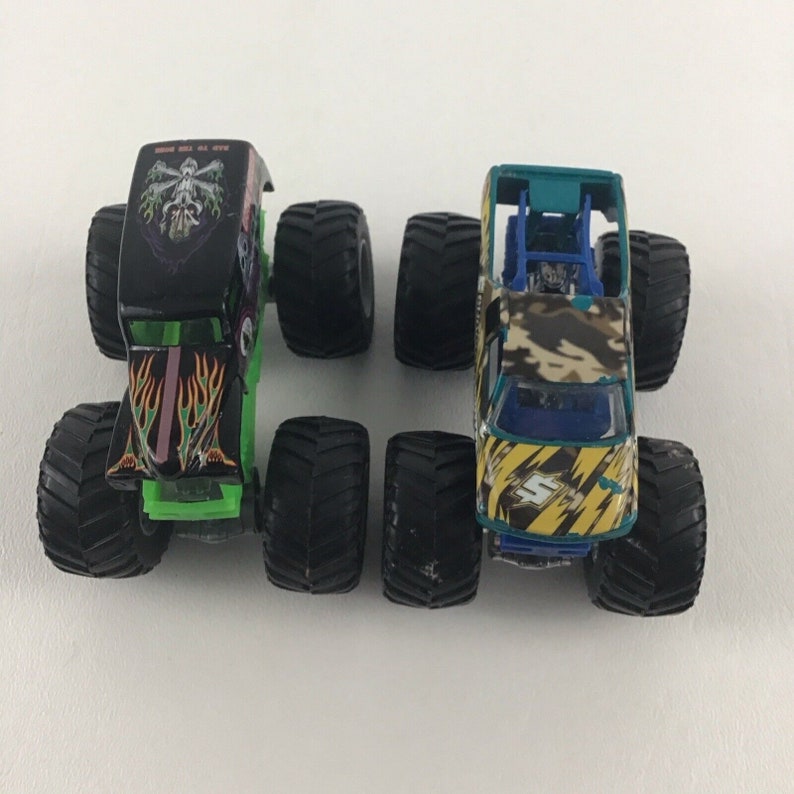 May include: Two monster truck toys. One is black with green flames and a skull on the hood. The other is yellow and green with a camouflage pattern and a blue windshield.