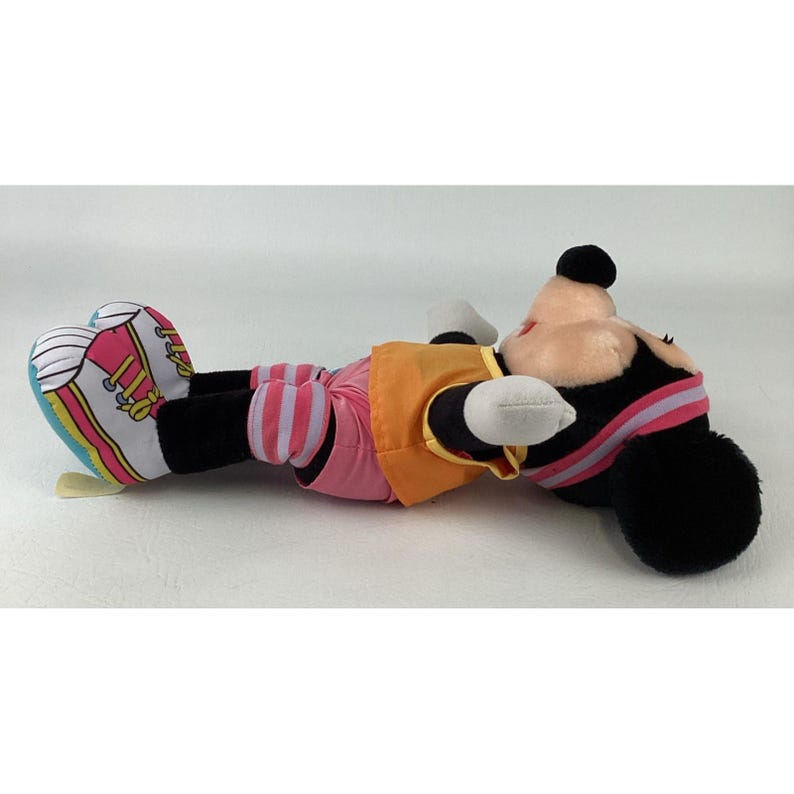 May include: A plush toy of a character with black ears, a tan face, and a black nose. The toy wears an orange vest, pink shorts with white stripes, and white sneakers with pink and yellow accents. The character has a pink headband with a white stripe.