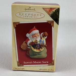 Könnte beinhalten: Hallmark Keepsake Weihnachtsschmuck in einer Box. Der Schmuck zeigt den Weihnachtsmann mit einer Glocke und einem Sack voller Spielzeug. Auf der Box steht "Santa's Magic Sack" und "Christmas 2005".