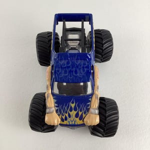May include: A blue toy monster truck with large black tires. The truck has a roll cage and is decorated with yellow and blue flame graphics. The arms of a figure are attached to the sides of the truck.