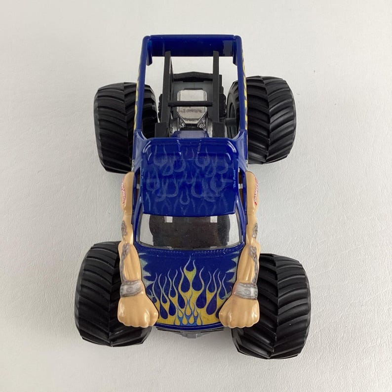 May include: A blue toy monster truck with large black tires. The truck has a roll cage and is decorated with yellow and blue flame graphics. The arms of a figure are attached to the sides of the truck.