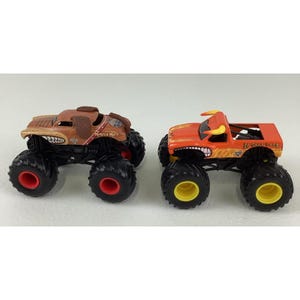 May include: Two toy monster trucks. One is brown with a dog-like design and the words "Monster Mutt". The other is orange with a bull design and the words "El Toro Loco". Both have large black tyres with red and yellow accents.