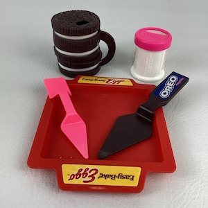 Op de afbeelding: Een verzameling speelgoedkeukenartikelen, waaronder een rode Easy-Bake Eggo-pan, een roze spatel, een zwarte Oreo-spatel, een witte container met een roze deksel en Oreo-koekjesvormige mokken.