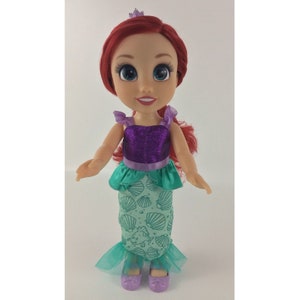 May include: A red-haired doll wearing a purple and green mermaid costume with a crown. The doll has blue eyes and a smile.