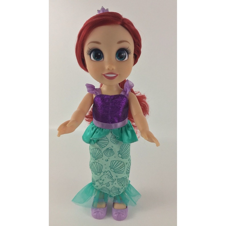 May include: A red-haired doll wearing a purple and green mermaid costume with a crown. The doll has blue eyes and a smile.