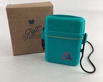 Pode incluir: Uma caixa retangular Avon Gift Collection de cor verde-azulado com um recipiente a condizer. O recipiente tem uma faixa amarela e um cordão preto. A caixa é castanha com as palavras "Gift Collection" impressas.