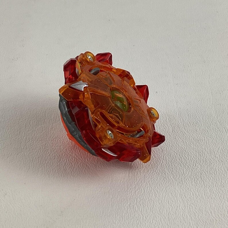 May include: A red and orange toy with a complex design. The toy has a transparent orange top with a central green element and is surrounded by red and gray components. It appears to be a spinning top or similar game piece.