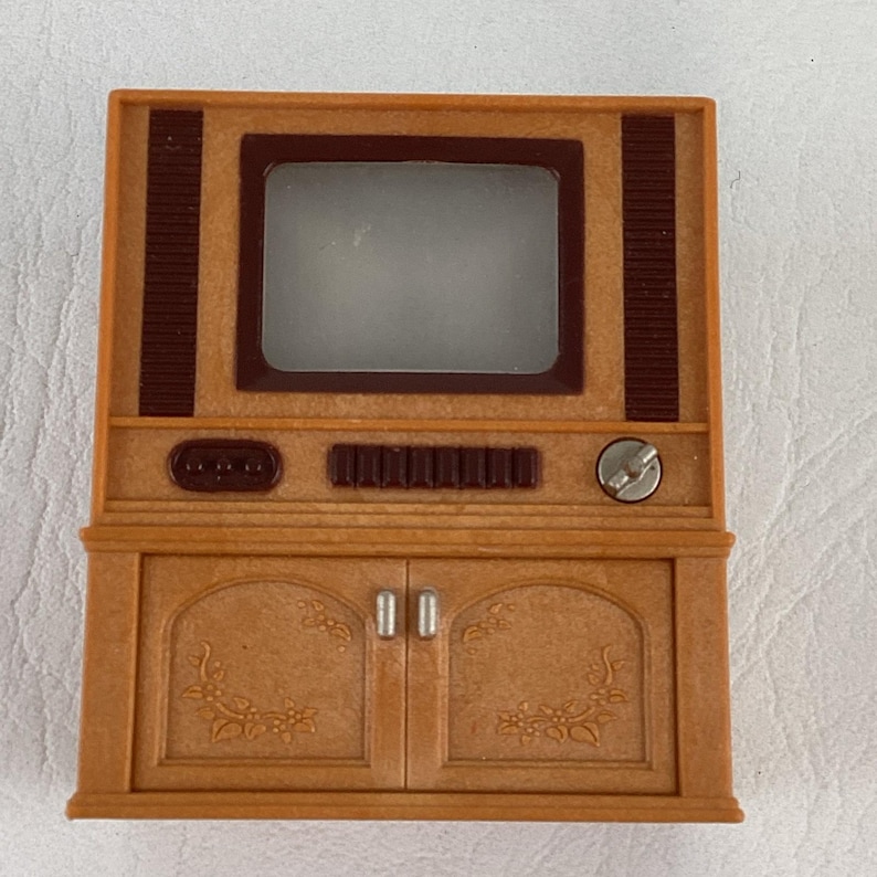 Op de afbeelding: Een miniatuur tv-toestel in vintage-stijl in bruin- en beigetinten. De tv heeft een rechthoekig scherm, een bedieningspaneel en een kast met twee deuren. Het ontwerp bevat decoratieve bloemmotieven.