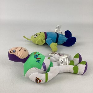 Pu&ograve; includere: Due peluche raffiguranti un personaggio ranger spaziale in una tuta bianca, verde e viola e un alieno verde a tre occhi in una tuta blu. Entrambi i giocattoli hanno etichette in tessuto bianco con testo stampato e sono appoggiati.
