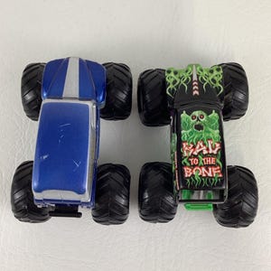 May include: Two toy monster trucks. One is blue with a silver stripe, the other is black with a green monster graphic and the text "RAV TO THE BONE". Both have large black tires.