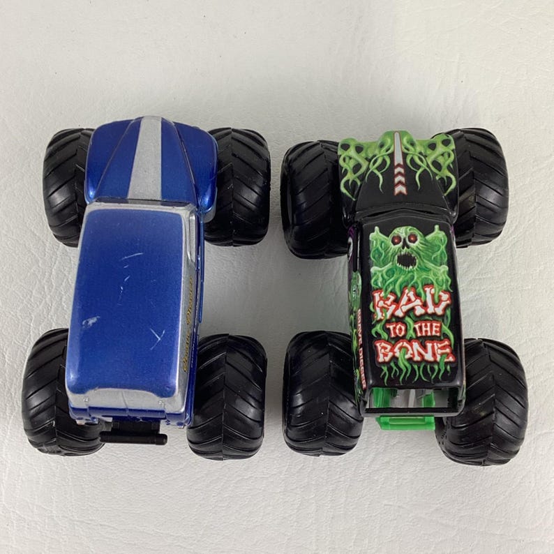 May include: Two toy monster trucks. One is blue with a silver stripe, the other is black with a green monster graphic and the text "RAV TO THE BONE". Both have large black tires.