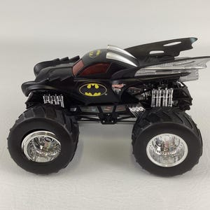 May include: Black monster truck toy with large tires and chrome rims. The truck features a Batman logo and wing-like extensions. The cab has a red tinted window. The toy is designed with a detailed suspension system.