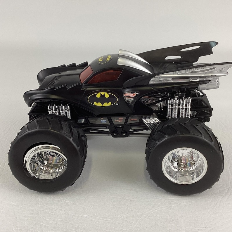 May include: Black monster truck toy with large tires and chrome rims. The truck features a Batman logo and wing-like extensions. The cab has a red tinted window. The toy is designed with a detailed suspension system.