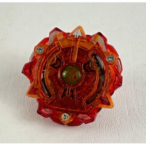 May include: A red and orange toy with a circular design. The toy has a central green circle with a letter inside, surrounded by intricate patterns and three silver screws. The outer edge has pointed, angular shapes.