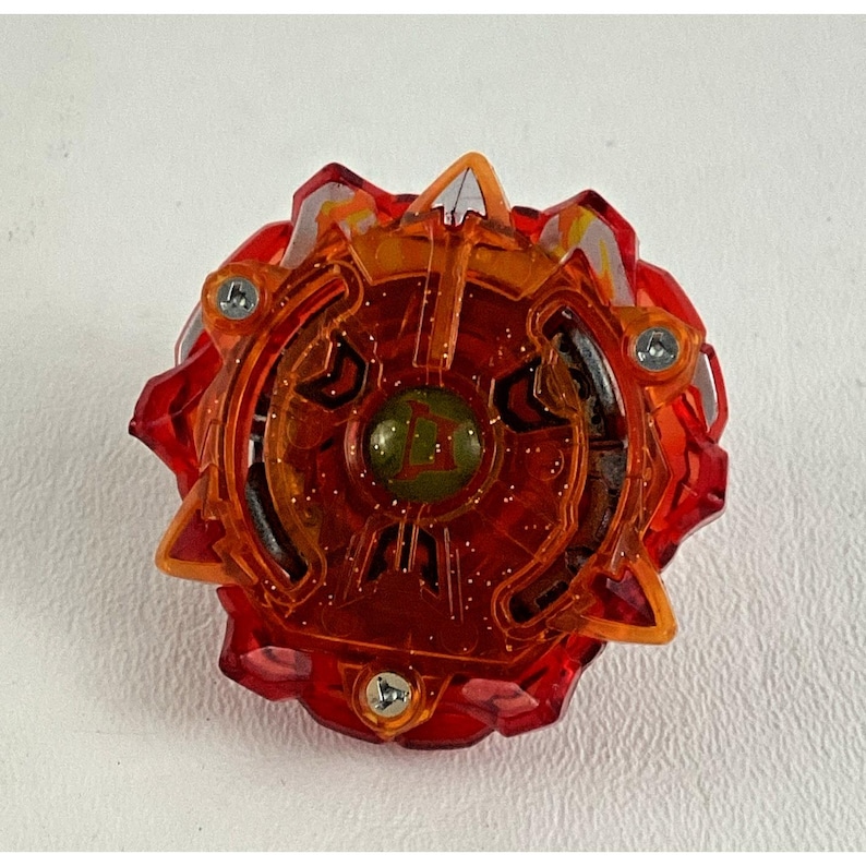 May include: A red and orange toy with a circular design. The toy has a central green circle with a letter inside, surrounded by intricate patterns and three silver screws. The outer edge has pointed, angular shapes.
