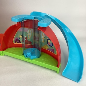Pu&ograve; includere: Un colorato playset in plastica con una struttura a casa rossa e verde, un ascensore trasparente e uno scivolo blu. Il playset presenta dettagli di personaggi dei cartoni animati ed &egrave; progettato per il gioco dei bambini.