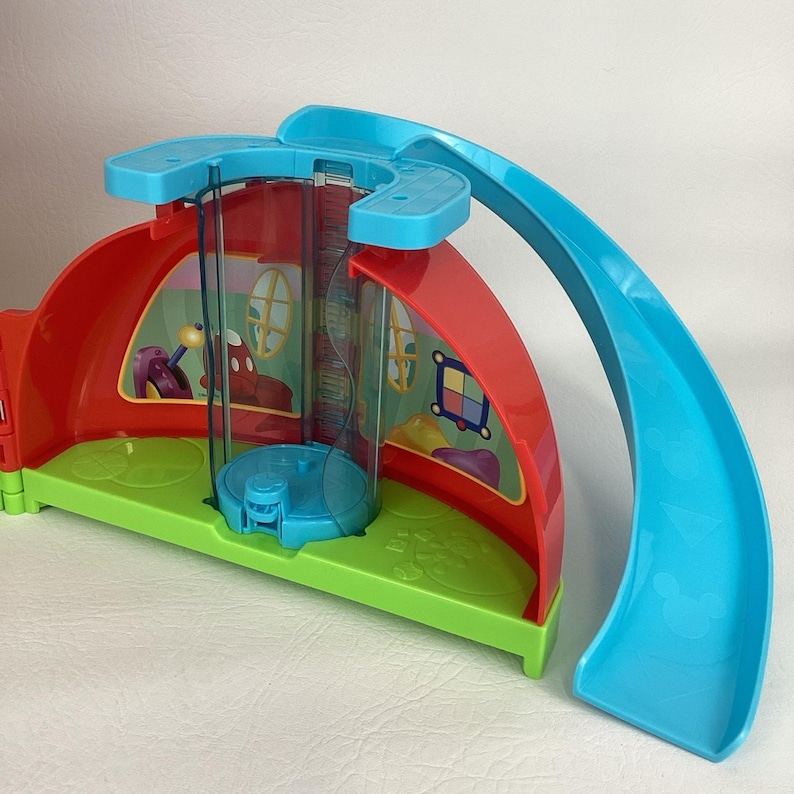 Pu&ograve; includere: Un colorato playset in plastica con una struttura a casa rossa e verde, un ascensore trasparente e uno scivolo blu. Il playset presenta dettagli di personaggi dei cartoni animati ed &egrave; progettato per il gioco dei bambini.