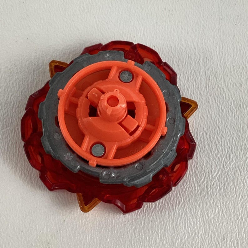 May include: A red and orange toy top with a gray outer ring. The top has a multi-layered design with a central orange component and a translucent red outer layer. The gray ring has a series of small, pointed protrusions.