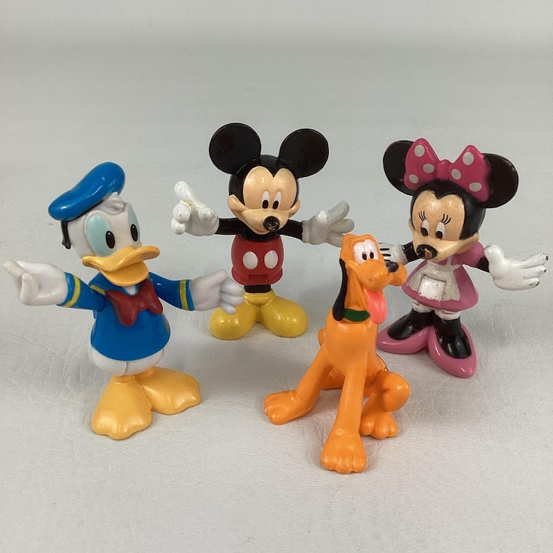 Pu&ograve; includere: Un gruppo di statuette dei personaggi Disney: Topolino, Minnie, Paperino e Pluto. Topolino indossa abiti rossi e neri, Minnie un vestito rosa, Paperino una tuta blu da marinaio e Pluto &egrave; arancione.