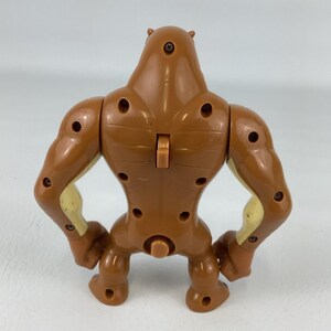 May include: A brown plastic action figure viewed from the back, featuring a muscular build with visible screw holes, a central vertical switch on the upper back, and light tan accents on the inner arms and torso sides, standing on two feet.