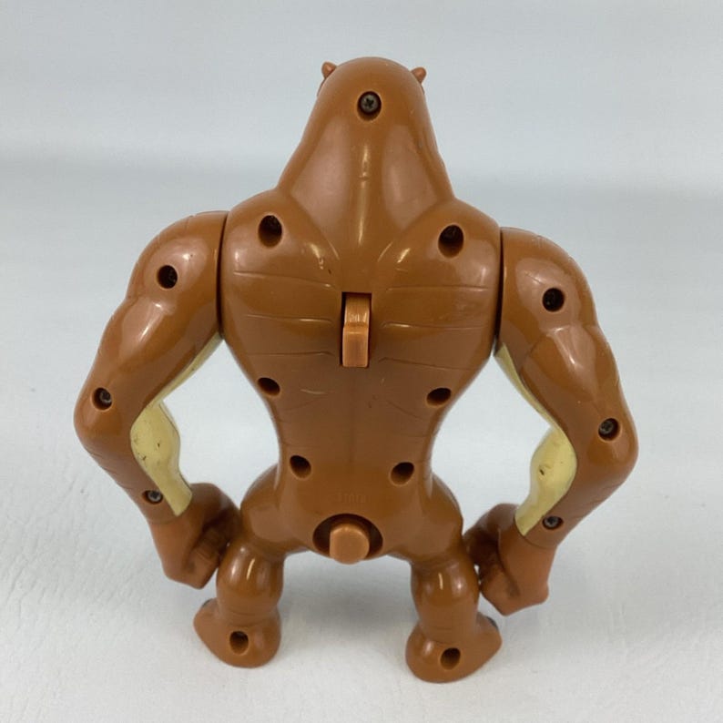 May include: A brown plastic action figure viewed from the back, featuring a muscular build with visible screw holes, a central vertical switch on the upper back, and light tan accents on the inner arms and torso sides, standing on two feet.
