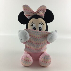 Op de afbeelding: Roze en zwarte pluche Minnie Mouse speelgoed met een roze stippen strik en een roze stippen deken. Het speelgoed zit en houdt de deken voor zijn gezicht.