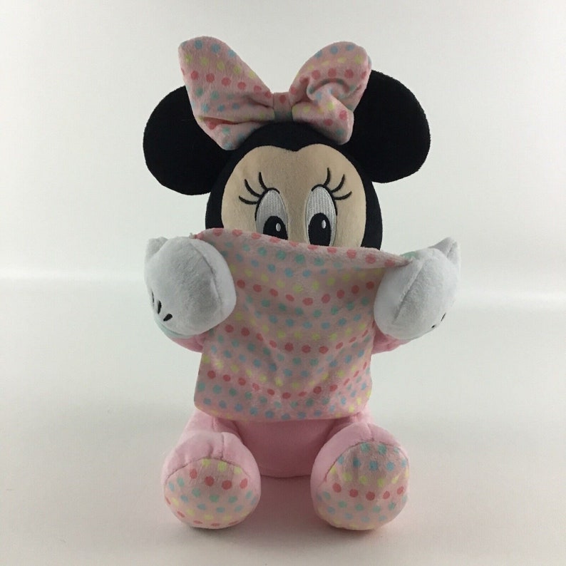 Op de afbeelding: Roze en zwarte pluche Minnie Mouse speelgoed met een roze stippen strik en een roze stippen deken. Het speelgoed zit en houdt de deken voor zijn gezicht.
