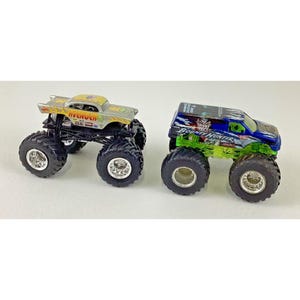 K&ouml;nnte beinhalten: Zwei Spielzeug-Monstertrucks. Einer ist silber und gelb mit dem Wort "Avenger" an der Seite. Der andere ist blau und gr&uuml;n mit den Worten "Bounty Hunter". Beide haben gro&szlig;e schwarze Reifen.