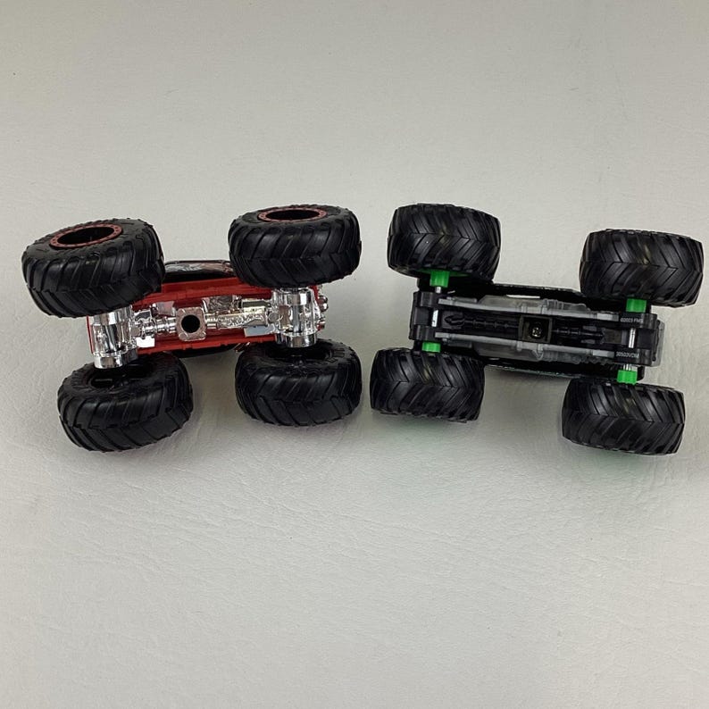 May include: Two toy monster trucks. One is red with black tires and a silver chassis. The other is black with black tires and green suspension components. Both have large, knobby tires.