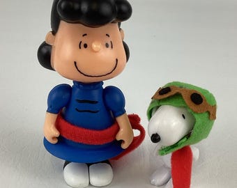May include: A pair of Peanuts figurines. One is Lucy in a blue dress with a red sash. The other is Snoopy wearing a green pilot's cap and a red scarf. Both figures are standing on a white surface.