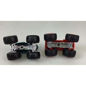 Pu&ograve; includere: Due monster truck giocattolo, uno turchese e argento, l'altro rosso. Entrambi hanno grandi pneumatici neri con cerchi rossi. I camion sono capovolti, mostrando i loro sottocarri e i battistrada dei pneumatici. Un giocattolo divertente per bambini.