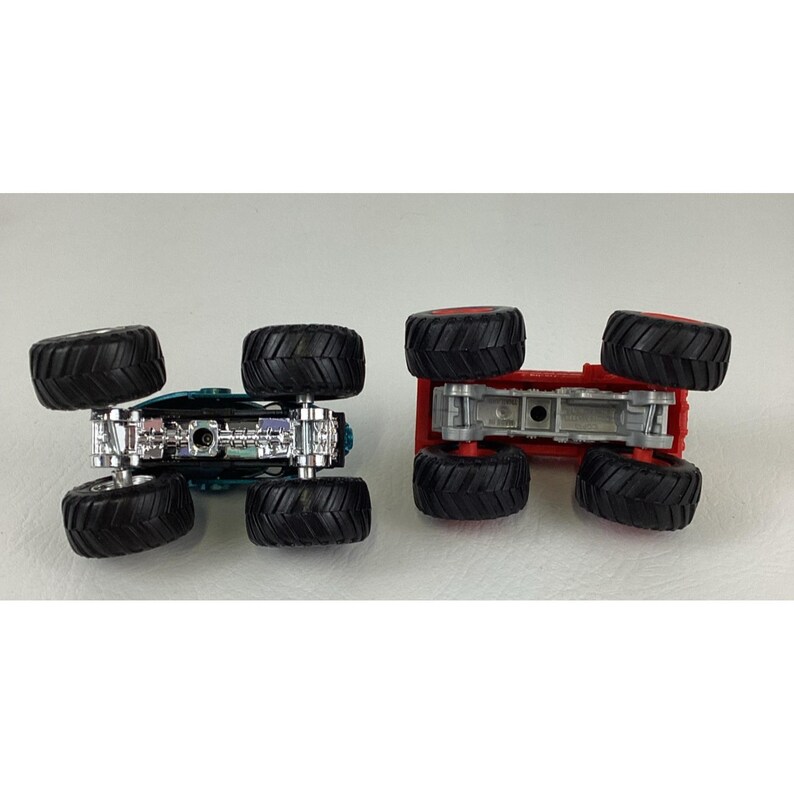 Pu&ograve; includere: Due monster truck giocattolo, uno turchese e argento, l'altro rosso. Entrambi hanno grandi pneumatici neri con cerchi rossi. I camion sono capovolti, mostrando i loro sottocarri e i battistrada dei pneumatici. Un giocattolo divertente per bambini.