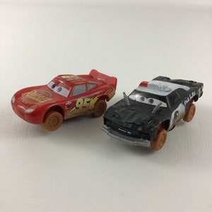 May include: Two toy cars from the Disney Pixar Cars movie franchise. The red car is Lightning McQueen, with the number 95 on the side. The black car is a police car with a white and black paint scheme. Both cars have a worn, dirt-covered look.