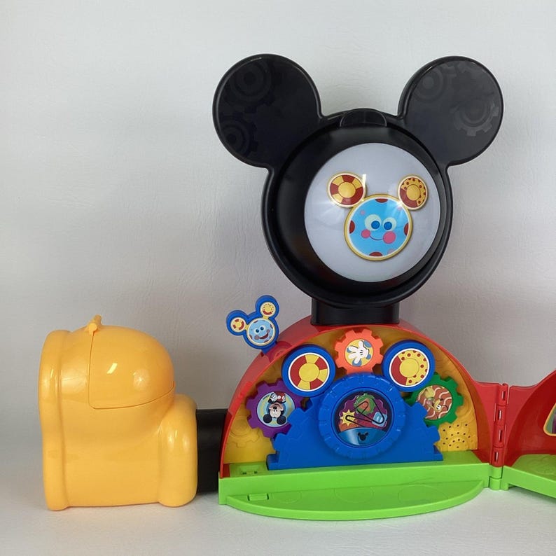Pu&ograve; includere: Un colorato giocattolo di Mickey Mouse Clubhouse con una grande testa e orecchie nere, un viso rotondo con un personaggio sorridente e vari ingranaggi. Il giocattolo &egrave; principalmente rosso, giallo, blu e verde.