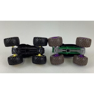 May include: Two toy monster trucks, one black with yellow accents and the other green with purple accents. Both have large, textured tires and are designed for off-road play. The trucks are shown upside down.