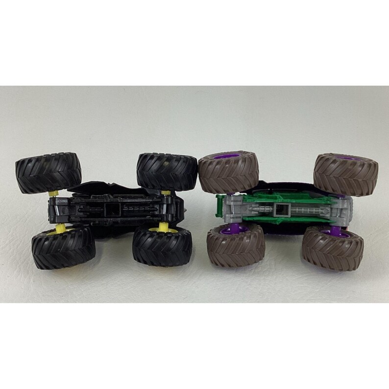 May include: Two toy monster trucks, one black with yellow accents and the other green with purple accents. Both have large, textured tires and are designed for off-road play. The trucks are shown upside down.