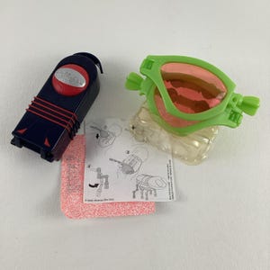 Könnte beinhalten: Ein grünes Plastikspielzeug mit einer roten und schwarzen Spielzeugfernbedienung. Das Spielzeug ist in Form einer Brille und hat eine durchsichtige Plastikbasis. Die Fernbedienung hat einen roten Knopf und einen schwarzen Griff. Das Spielzeug ist mit "Miramax Films Corp." beschriftet.