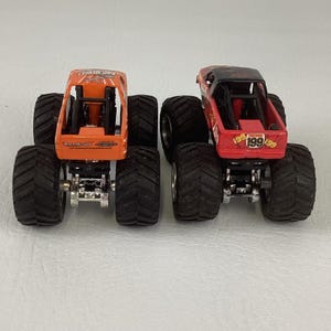 Puede incluir: Dos camiones monstruo de juguete, uno naranja y otro rojo, con neum&aacute;ticos negros grandes. El cami&oacute;n naranja tiene el texto "BAD NEWS!" y "Race Wraps". El cami&oacute;n rojo tiene el n&uacute;mero "199" en la parte trasera.