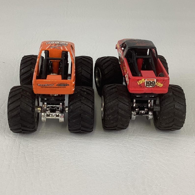 Puede incluir: Dos camiones monstruo de juguete, uno naranja y otro rojo, con neum&aacute;ticos negros grandes. El cami&oacute;n naranja tiene el texto "BAD NEWS!" y "Race Wraps". El cami&oacute;n rojo tiene el n&uacute;mero "199" en la parte trasera.