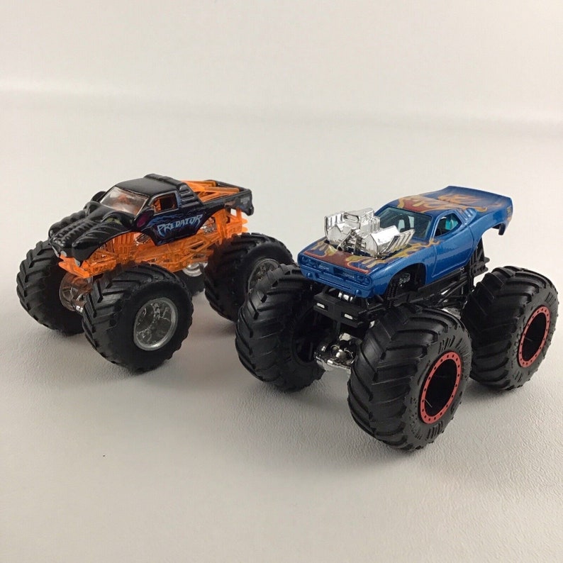 May include: Two monster trucks, one black with orange accents and the word "Predator" on the side, and one blue with orange flames and a silver engine. Both trucks have large black wheels.