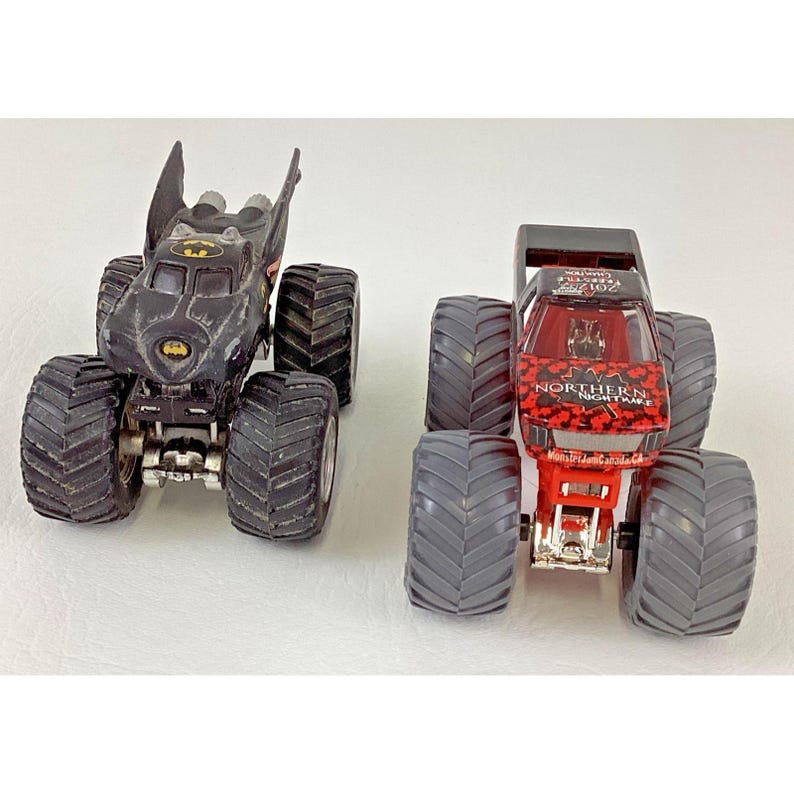 May include: Two toy monster trucks. One is black with a Batman logo and bat wings. The other is red and black with the text "Northern Nightmare". Both have large, gray tires.