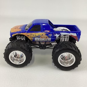 Op de afbeelding: Een blauwe en oranje speelgoed monstertruck met grote zwarte banden en chromen velgen. De truck heeft de tekst "KING KRUNCH" en een afbeelding van een gespierde man. De laadbak heeft een blauw en wit sterrenontwerp.
