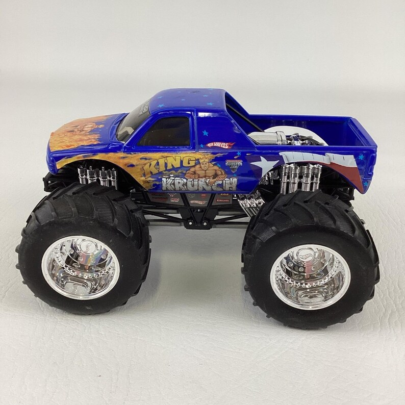 Op de afbeelding: Een blauwe en oranje speelgoed monstertruck met grote zwarte banden en chromen velgen. De truck heeft de tekst "KING KRUNCH" en een afbeelding van een gespierde man. De laadbak heeft een blauw en wit sterrenontwerp.